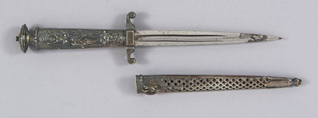 Petite dague de bureau. Manche en laiton ciselé argenté à décor du diable. Lame à deux tranchants. Fourreau en laiton ciselé travail...